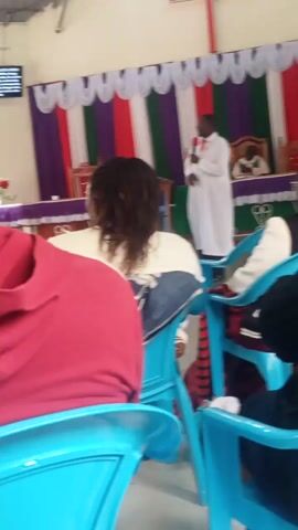 A religious service is taking place in a room that appears to be a church. A man in a white robe and a purple sash is standing on a raised platform and appears to be speaking or preaching. He is holding a microphone. Behind him, there is a wall decorated with colorful striped fabric and white ruffled trim. Several people are seated in blue plastic chairs, facing the platform. The camera is positioned behind some of the seated individuals, showing their backs and heads. One person in the foreground is wearing a red top. The room is lit, and the overall atmosphere suggests a gathering for worship.