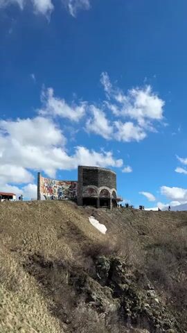 The video displays a structure atop a hill against a blue sky with scattered white clouds. The structure features a curved mural with multiple figures and a stone tower with arched openings. Several people are seen around the structure. A patch of snow is visible near the base of the structure. The hill is covered in dry vegetation and rocky outcrops. In the background, a snow-covered mountain range stretches across the horizon. The camera gradually pans right, revealing more of the mountain range.