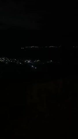 A blurry, dark video shows a night landscape with scattered lights in the distance, suggesting a city or town. The camera appears to be moving, causing further blurring. No people or specific objects are clearly visible due to the low light and motion. The environment is dark, with the only illumination coming from artificial lights.