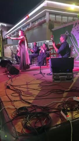 A female singer in a long, dark dress with a patterned scarf stands center stage, holding a microphone and gesturing with her hands. Behind her, three other performers are seated. A man on the left plays a keyboard, another man in the center claps his hands, and a woman to his right also claps. To the right of the seated performers, a man plays a guitar. The stage is lit with pink and red lights, and numerous black cables are visible on the stage floor in the foreground. In the background, a building with illuminated windows and decorative plants is visible. The scene appears to be an outdoor performance at night.