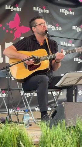 A man with dark hair and glasses is sitting on a chair playing an acoustic guitar and singing into a microphone. He is wearing a dark short-sleeved shirt and dark pants. The guitar is light brown and has a pickguard. A music stand with a sheet of music is in front of him. He is performing on a stage with a black backdrop that has the word "BAKgarden" and a pink butterfly logo repeated. There is green grass in the foreground.