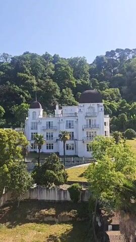A static, high-angle, outdoor shot captures a large, white building with two domed towers, balconies, and multiple windows. The building is situated on a lush green hillside, surrounded by trees and palm trees under a clear blue sky.