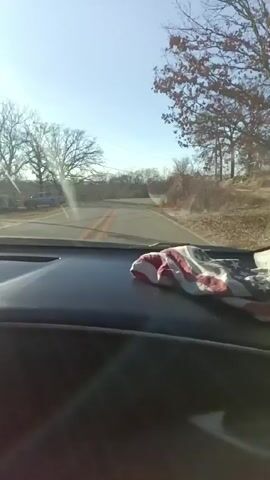 The video is taken from inside a car driving along a narrow country road. The weather appears to be sunny with a clear blue sky. The trees lining the road are mostly bare, indicating it may be late fall or winter. An American flag themed cloth is draped over the dashboard. There are utility poles and wires visible above the road. In the distance, the road curves slightly. The dashboard and interior of the car appear dark, contrasting with the bright outdoor scene. Music is playing in the background.