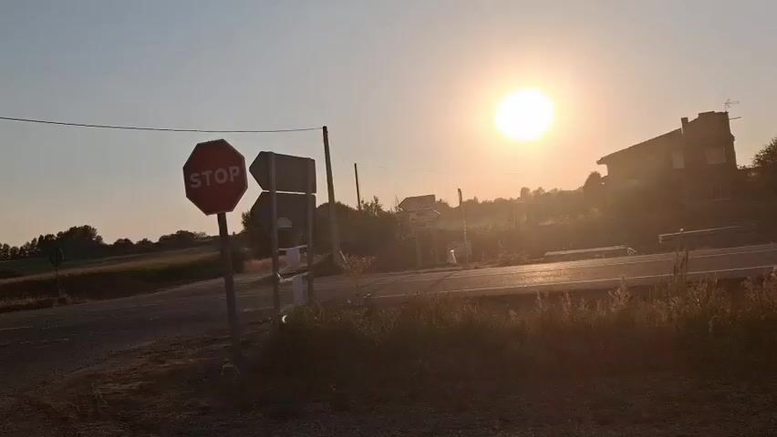 The video opens with a view of a road intersection at sunrise or sunset. A stop sign is visible on the left, along with directional signs pointing to different towns. The sun is low in the sky, casting a warm glow over the scene. The landscape appears rural, with fields and trees in the background, and a house is visible on the right. As the video progresses, the camera focuses on the sky, revealing a partial solar eclipse. The sun is partially obscured by the moon, creating a crescent shape. The light in the sky changes from a bright yellow to a hazy orange. The video concludes with a close-up view of the obscured sun.