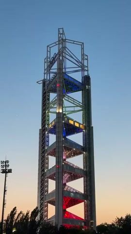 The video shows a tall, modern tower-like structure illuminated with colorful lights, mostly red, blue, green, and yellow. The structure has an open framework design with several platforms. The sky in the background transitions from orange near the horizon to blue above. To the left of the structure is a tall light pole with multiple lights. Some trees are visible at the bottom of the frame. The tower's lights change color throughout the video.
