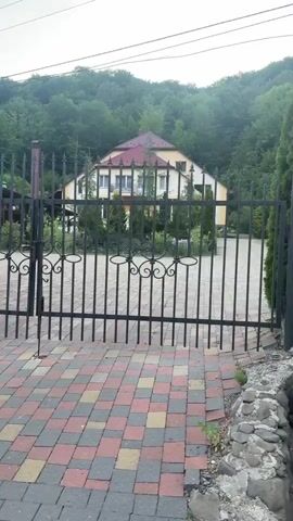 The camera slowly pans right, revealing a house and a decorative wrought-iron gate and fence. The house, visible through the gate, has a light-colored exterior and a red roof. Behind the house is a green, wooded hill. In front of the gate, the ground is covered with multi-colored pavers. As the camera pans right, it shows the fence line, which runs along a low rock wall. Behind the fence are tall, slender, evergreen trees that form a dense screen. Beyond the rock wall is a narrow grassy area and a road. The sky is overcast.