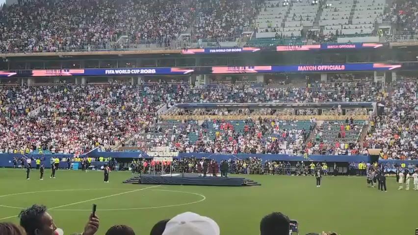 A panoramic view shows a packed football stadium filled with spectators. The field is green, with a raised platform in the center where several people in formal attire are gathered. Some individuals in dark uniforms appear to be security personnel. In the background, banners proclaiming "FIFA WORLD CUP 2026" and "BRONZE FINAL WINNERS" are visible. The atmosphere suggests a pre-game ceremony or awards presentation.