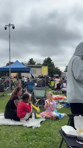 A group of people are gathered outdoors at an event. The scene is likely a park or festival, with grass covering the ground. Several blue tents and a stage are visible in the background, suggesting a performance or organized gathering is taking place. Many people are seated in lawn chairs or on blankets, some appear to be eating. In the foreground, a woman and two children are sitting on a blanket. One child, a girl wearing a purple top and a colorful skirt, is eating a hot dog. Another child, a boy wearing a red sweatshirt, is also eating. Behind them, more people are visible sitting in chairs. The sky is overcast with clouds. There is a CCTV camera mounted on a tall pole in the upper left portion of the frame.