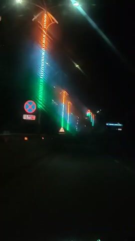 The video shows a night-time drive down a dark road. On the left side of the road, there are streetlights decorated with vertical LED lights in green, white, and orange, resembling the Indian flag. A red and blue circular sign with a white diagonal bar and a red border is also visible on the left. Further down the road, more of these illuminated streetlights are visible. A triangular yellow warning sign with two arrows indicating a split in the road is also present. The overall impression is a festive or patriotic display of lights along the roadside during nighttime.