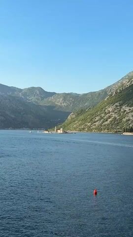 The video shows a wide shot of a bay with mountains in the background. In the middle of the water, there is a small island with a church. The sky is clear and blue, and the water is calm and blue. The island appears to be man-made, possibly with a dock or pier. The mountains are covered in green vegetation. There is a red buoy in the foreground of the water. The scene suggests a peaceful, scenic location.