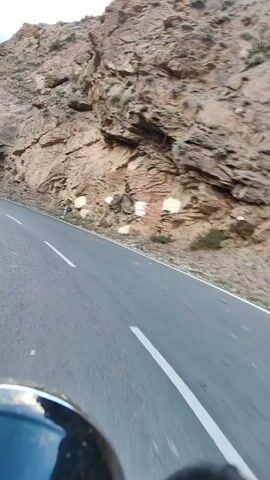 This video captures a first-person perspective of a motorcycle ride on a winding mountain road in Stingri, India. The scene is dominated by a steep, rocky mountainside on the right, with a dramatic drop-off to a valley on the left. The road is paved and well-maintained, marked with white dashed lines.

The time of day appears to be late afternoon or early evening, as suggested by the soft, warm light illuminating the mountain peaks and the shadows cast across the landscape. The weather seems clear and pleasant, conducive to riding.

In the background, lush green slopes and more rugged mountain ranges are visible. A few structures and signs can be spotted on the left side of the road, hinting at a small settlement or a service area. The rider's helmet and handlebars are visible in the foreground, indicating the point of view.

The overall mood is one of adventure and exploration, with the vastness of the mountains and the thrill of navigating the serpentine road creating a sense of freedom. The video is a visual journey through a stunning natural landscape, highlighting the beauty of the Indian Himalayas. No discernible text is visible within the frame.