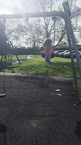 A young girl with brown hair and glasses is swinging back and forth on a tire swing in a park. She is wearing a pink t-shirt and purple pants. The sun is shining brightly, and there are trees and cars in the background. The girl is smiling and appears to be enjoying herself. The tire swing is attached to a metal frame. The ground beneath the swing is covered in wood chips. There is another swing set in the background.