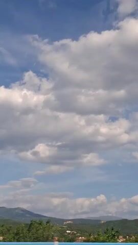 The video shows a view from a vehicle moving through a rural landscape. The sky is partly cloudy with a mix of white and gray clouds against a blue sky. Below the sky is a green, hilly landscape dotted with trees and some buildings. The trees in the foreground appear blurred due to the motion of the vehicle. There are no people or animals visible in the video. The overall impression is a scenic drive through the countryside on a pleasant day.