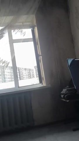 The video begins by showing a window with sheer curtains blowing in the wind, revealing apartment buildings outside. The camera pans to the right, showing a sofa with blankets on it, and then to a child standing in front of the sofa. The child is wearing a black t-shirt and grey shorts, and is barefoot. The child looks at the camera and smiles. There is a power bank and charging cable on the floor next to the sofa.