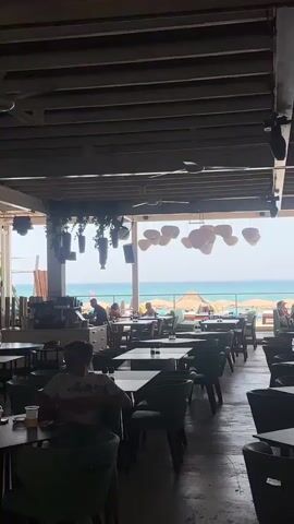 The video pans across a restaurant with a view of the ocean. Tables and chairs are set up inside and outside. Several people are seated at tables, and some are walking around. A DJ booth is visible near a glass enclosure. The restaurant appears to be open for business, with patrons enjoying the beachfront atmosphere.