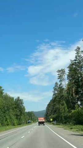 The video shows a front-facing view from a vehicle driving on a two-lane asphalt road. An orange dump truck is visible ahead, traveling in the same direction. The road is bordered by dense green trees on both sides, with the right side featuring taller, coniferous trees and the left side a mix of deciduous and coniferous trees. In the distance, faint mountain or hill outlines are visible under a clear, bright blue sky with scattered white clouds. A yellow, diamond-shaped road sign with a black pedestrian crossing symbol is visible on the right side of the road. The weather appears sunny and clear. The setting suggests a rural or forested area, possibly near mountains, in Souzga, Russia. The overall scene conveys a sense of travel through a natural landscape.