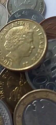 The video is a close-up, static shot of a pile of various coins. The focus is on a gold-colored coin with the profile of Queen Elizabeth II and the year 2013 visible. Behind it, other coins are visible, including a silver-colored coin with the word "ELIZABETH" and a gold-colored coin with what appears to be a dragon. The coins are piled on top of each other in a shallow depth of field, obscuring any background details. The lighting is bright and appears to be artificial.
