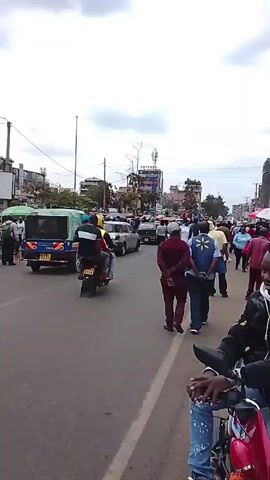 The video shows a busy street in a city, likely in Africa. Many people are walking on the sidewalk and along the side of the road, some congregating. There are cars, motorcycles, and a tuk-tuk moving through the street.  Buildings line the street, with various businesses and signs visible. The sky is cloudy and overcast. The camera appears to be handheld and stationary, taken from the perspective of someone on a motorcycle parked on the side of the road.