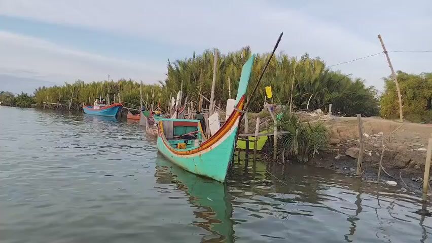 The video shows a tranquil river scene with several traditional fishing boats docked along the bank. The water is calm, reflecting the blue sky and the lush greenery of palm trees lining the river. One boat, painted in a vibrant turquoise color with red and orange accents, is prominently featured in the foreground. The boat has a tall, curved bow and is tied to a wooden pole driven into the muddy bank. Other boats, in various colors including blue and red, are visible further down the river, also moored along the shore. The overall atmosphere is peaceful and idyllic, suggesting a quiet moment in a coastal or riverside community.