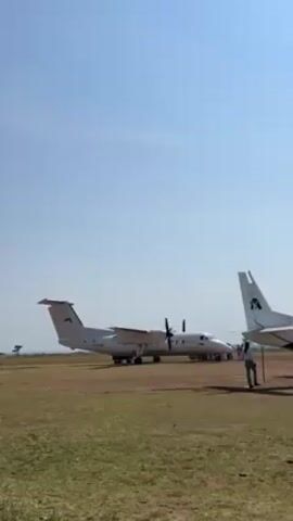 The video shows a white turboprop airplane on a dirt runway. Another airplane is partially visible on the right side of the frame. The sky is clear and blue. A person is standing next to the plane, holding a long, thin object that appears to be a stick. The propellers are spinning, creating a loud whirring sound. The environment looks like a rural airstrip in Africa.