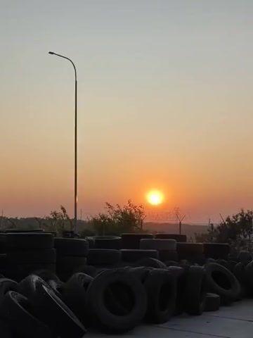 The video shows an outdoor scene at sunset. The sun is visible as a bright orb in the orange sky. In the foreground, there is a large pile of used tires. Behind the tires, there is a barbed wire fence and some trees. A single lamppost stands tall on the left side of the frame. The overall impression is one of a desolate industrial or junkyard setting bathed in the warm light of dusk.