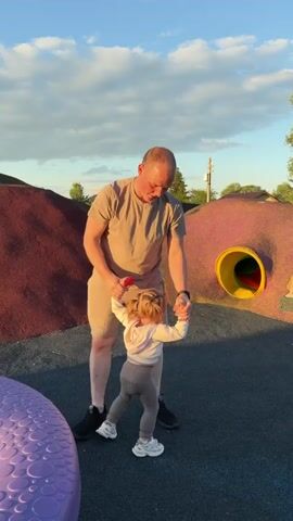 Here's a description of the video:

The video shows a man and a toddler at a playground on a sunny day. The man is fair-skinned, wearing a beige t-shirt and shorts with dark sneakers. He's standing near a toddler, who has blonde hair in pigtails and is wearing a white hooded sweatshirt and gray leggings with white sneakers. They are on a soft, dark gray playground surface.

The playground has several colorful structures, including a large red and purple dome-shaped mound and another dome covered in red and speckled pink. This second dome has a yellow tunnel running through it. The toddler walks towards the tunnel with a red object in her hand. The man watches her, at one point raising his arm to scratch his head. In the background, there are trees, a house, and a few people on another part of the playground.