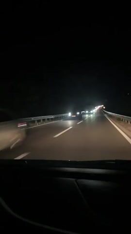 The video shows the view from the driver's seat of a car driving on a highway at night. Other cars are visible in front, with their headlights illuminating the road. The road markings are clearly visible. Guardrails line the edge of the highway. A road sign is also visible. It's dark, but the car's headlights and those of other vehicles provide some visibility. The camera is stable, and the video appears to be shot from inside the car. The overall impression is a nighttime drive on a busy highway.