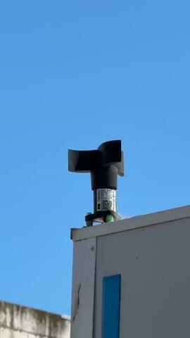 This is a short video clip showing a close-up of a black anemometer mounted on top of a white, outdoor electronic unit. The anemometer has two curved blades that are positioned to catch the wind. It is attached to a cylindrical housing that has a label with text and a QR code. The unit itself is positioned on the edge of a flat rooftop, with a clear blue sky in the background. To the left and slightly below the unit, a white brick wall is partially visible. A rectangular blue panel is also visible on the side of the white unit. The lighting suggests it is daytime, likely with sunny weather. The camera angle is slightly upwards, focusing on the anemometer and the top of the unit. The video is static, with no movement of the camera. There is no discernible activity taking place other than the potential movement of the anemometer blades due to wind. The label on the anemometer contains a barcode and text, but it is not legible enough to read.
