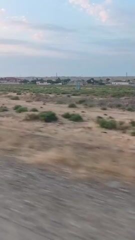 The video shows a blurry, outdoor scene from the perspective of a moving vehicle. The foreground is dominated by a gray, textured surface that appears to be a road or dirt path. Beyond this, the landscape is dry and arid, with patches of brown, yellow, and green vegetation. In the distance, there are several small buildings, some with visible roofs and structures. A tall, blue water tower stands out among the buildings. The sky is partly cloudy with a pale blue hue, suggesting it might be early morning or late afternoon. The overall impression is of a rural or semi-rural area with a somewhat desolate or undeveloped appearance. The movement of the vehicle causes the scene to be out of focus, particularly in the foreground.