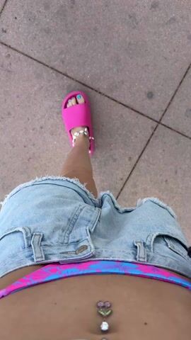 The video shows a person walking on a paved surface. The person is wearing denim shorts, a pink and blue swimsuit bottom, a belly button ring with a flower design, and pink sandals. The person has a tattoo on their leg. The ground is a light pinkish-gray with dark gray lines, possibly indicating tiles or slabs. The camera perspective is from directly above, looking down at the person's lower body and feet as they move forward.