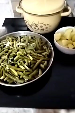 A top-down video shows a cooking scene. In the foreground, a large, shallow, grey pan is filled with chopped green beans, some of which appear to be blanched or lightly cooked. To the right of the pan, a small white bowl contains cubed, raw potatoes. In the background, a large, cream-colored pot with a floral pattern sits on a stovetop. The stovetop is black. The overall impression is of ingredients being prepared for a meal.