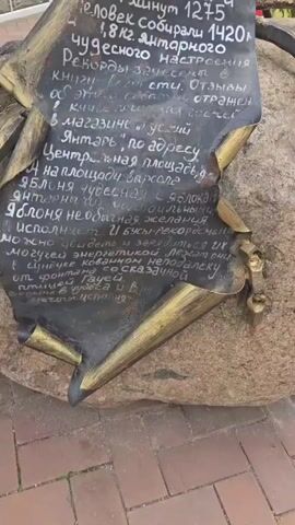 This video begins with a close-up of a metal plaque attached to a large rock, inscribed with Cyrillic text and what appears to be decorative golden elements. The plaque details an event that occurred on July 1, 2017, involving a "miracle," a duration of 4 hours and 12 minutes, and the collection of 1420 pieces of amber weighing 1.8 kilograms. It also mentions records, a book of achievements, reviews, and mentions a store at a specific address on a central square, near an apple-themed landmark. The camera then pans up, revealing a metallic sculpture of a tree with amber-like fruits attached to its branches. In the background, a building with the word "КОФЕЙНЯ" (which translates to "Coffee Shop") is visible, along with a red vintage car and more of the building's facade. The video continues to pan upwards, showing more of the tree sculpture against the sky and the building.