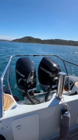 The video captures a serene daytime scene from the stern of a boat, looking out over clear, blue-green water towards a distant coastline. The sky is bright blue with a few scattered white clouds, suggesting good weather conditions.

In the foreground, the back of the boat is visible. Two black Mercury outboard engines are mounted on the transom, both clearly displaying the "MERCURY" logo in white. The engines are running, creating a gentle hum and stirring the water behind the boat. Polished metal railings extend from the sides of the boat, curving around the stern. Wooden accents are visible on the deck area to the left, and part of an inflatable boat tube is also seen on the left side. A white storage compartment or hatch is located on the stern, with the letters "CRC" faintly visible on its surface. A gray cylindrical object, possibly a fender or buoy, hangs from one of the metal railings on the right.

In the mid-ground, the ocean water stretches out, shimmering under the sunlight with small ripples and waves. The water's clarity allows for some visibility beneath the surface. In the background, a rugged, hilly coastline is visible, covered in sparse vegetation and rocks. There are no prominent buildings immediately visible on the closest land, but some structures might be present further along the distant shore. The overall impression is one of a calm and pleasant boat ride in a scenic coastal area.