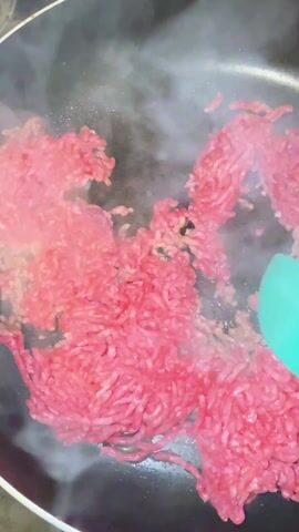 The video shows ground beef being cooked in a frying pan. A turquoise spatula is used to break up the ground beef as it cooks, causing steam to rise from the pan. Some pieces of the ground beef have already browned, while other portions remain pink. The pan appears to be non-stick and is placed on a stove.