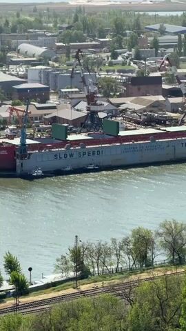 The scene is a high-angle, daytime shot of a port area. A large gray vessel is docked at the port, bearing the markings "SLOW SPEED!" and "ANCHORAGE PROHIBITED" in English and their Russian equivalents. The vessel is being serviced by multiple cranes. The surrounding area includes industrial buildings, storage tanks, and other docked ships. Below the port, a tree line is visible, with railroad tracks running alongside it. In the distance, a glimpse of the horizon is visible.