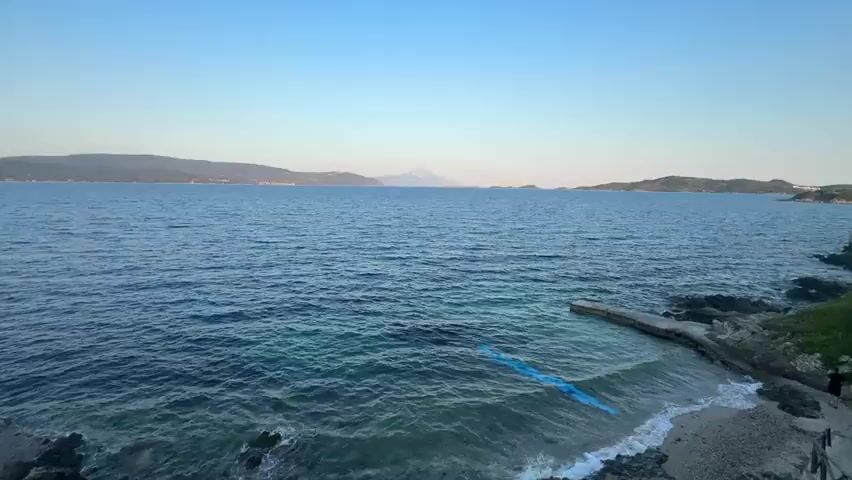 A drone view from a high angle shows a body of water and a shoreline. The water is deep blue and appears calm, with small waves lapping at the shore. There is a small concrete pier jutting out into the water. A person can be seen walking on the shoreline near the pier. The shoreline is rocky and covered with pebbles. The background shows a distant coastline with hills and mountains. The sky is clear and blue, with the sun beginning to set. A blue object, possibly a floating mat or raft, is visible in the water near the shore.