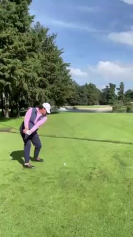 A person is seen playing golf. They are wearing a light pink long-sleeved shirt, a navy blue vest, dark pants, black shoes, and a white hat. They swing a golf club, hitting the ball. After the swing, they walk forward on the green golf course towards a water hazard with a small white barrier. The golf course is surrounded by lush green trees and a partially cloudy blue sky.