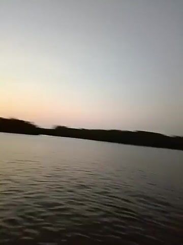 The video shows a view from a boat on a body of water. The water has small waves and reflects the sky. In the distance, there is a dark treeline. The sky is light blue with some clouds and a band of orange and yellow light on the horizon. The camera angle shifts to show a part of the boat's interior, with a light-colored ceiling and wooden trim. A woman is speaking, asking if "we will check there." Another person says "yes." She then says "Sprite."