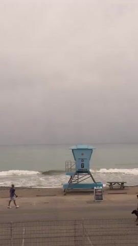 The video captures a serene, overcast beach scene, panning slowly from left to right. The ocean occupies the middle ground, its muted teal waters meeting a sandy and rocky shore with gentle white waves. The sky is a uniform, pale grey, indicating an overcast or foggy day.

In the foreground, a paved area, likely a parking lot, runs parallel to the beach. Initially, a silver SUV is parked on the left side of this paved area. A metal fence with a diamond mesh pattern separates the immediate foreground from the parking lot and beach.

As the camera pans, two individuals appear walking along the waterline on the beach, moving from left to right. One person wears dark clothing, while the other appears to be in red. Further along the paved area, several picnic tables and trash cans are visible. The pan continues to reveal a cluster of tall palm trees standing behind the paved area, adding a tropical touch to the coastal landscape. The overall mood is quiet and peaceful. There is no visible text in the video.