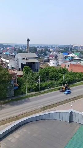 A drone shot shows a view of a town. A road runs through the middle of the frame, with trees and buildings on either side. A blue car and a white bus are driving on the road. A yellow bulldozer is also on the road. The buildings in the background include a tall, industrial-looking structure with a smokestack. The sky is clear and the sun is shining.