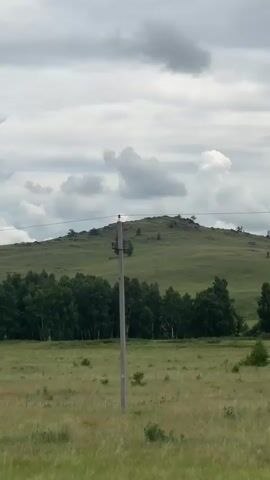 A time-lapse video shows a slightly cloudy sky above a grassy hill with a line of trees at its base. A single utility pole is visible in the foreground. The camera pans slowly from left to right across the landscape.