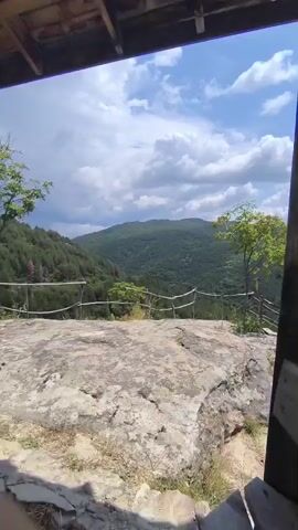 The video is a first-person perspective panning across a natural landscape. In the foreground, a wooden structure with a bench is visible. The camera moves to reveal a rocky outcrop with a rope railing, suggesting a viewpoint or observation deck. Beyond the railing, a vast expanse of rolling green hills and mountains unfolds under a bright blue sky dotted with white, fluffy clouds. Trees and vegetation cover the landscape, with some sparse dry grass in the immediate foreground. The overall scene depicts a scenic, natural environment on a clear day.