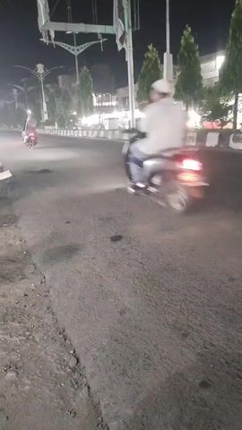 This is a shaky, low-light video shot from a low angle looking at a dark asphalt road at night. The road appears to be slightly damaged with some gravel and rough patches visible on the right side. On the left side of the road, a white motorcycle is parked with its back facing the camera. In the middle of the road, a person on a white scooter is riding away from the camera. Further down the road, several trees line the street with lights visible in the background, suggesting buildings or shops. A street light can be seen hanging over the road in the upper left corner of the frame. The scooter's brake light is illuminated, and the headlights of an approaching car can be seen in the distance, creating a hazy glow. Another vehicle, a dark-colored SUV, is shown driving past the scooter and away from the camera in the mid-ground, its headlights also visible. The overall atmosphere is that of a typical urban street scene at night with ongoing traffic.
