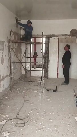 The video shows two men working in a room that is undergoing renovation. One man is on a metal scaffold applying plaster to a wall with a trowel. He is wearing a blue hooded jacket and dark pants. The second man is standing on the floor watching him. He is wearing a black outfit and a black beanie. The room appears to be in a state of disrepair, with exposed walls and debris on the floor. There is a doorway leading to another room. A drill is on the floor.