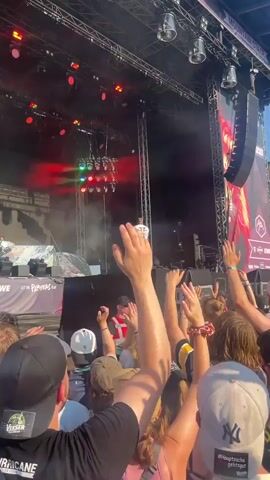 A live music performance is underway outdoors. A lone performer stands center stage, facing away from the audience, on a raised platform. Above the performer, a rig of bright red lights hangs suspended. The stage is equipped with large speakers mounted on either side of the structure. In front of the stage, a dense crowd of people are gathered, many with their hands raised in the air, appearing to be enjoying the show. The sky above is clear and blue, suggesting a daytime event. The overall atmosphere is energetic and celebratory.