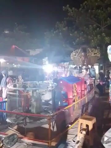 The video shows a busy outdoor scene at night with fairground rides. There is a circular ride with several children inside. A red and blue playhouse is located next to it, and a small stool is nearby. Several people are milling around, including adults and children. The scene is brightly lit by several lights. A red tent and trees are visible in the background. A motorcycle mirror is in the bottom left of the frame.