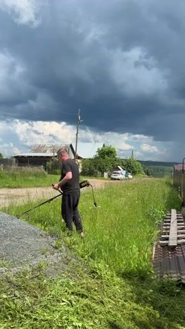 A man is trimming grass with a string trimmer in a rural setting. He is wearing a black t-shirt and black pants. He is standing in a grassy field next to a gravel path. In the background, there are houses, trees, and cars. The sky is cloudy and appears to be dark and stormy.