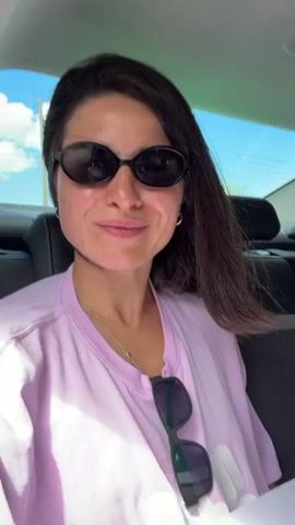 A woman with dark hair and sunglasses is filmed from a slightly elevated angle inside a car, likely in the backseat, in Bratešići, Montenegro. She is wearing a light pink t-shirt and a delicate silver necklace. Another pair of dark sunglasses is tucked into the collar of her shirt.

Throughout the video, she smiles and nods her head, seemingly enjoying music playing in the background. She looks around, sometimes directly at the camera, sometimes up, and at other times slightly off to the side. The car's interior is visible, including a headrest and parts of the seats. Through the car windows, a clear blue sky with some white clouds is visible, indicating a bright, sunny daytime setting. She appears relaxed and cheerful.