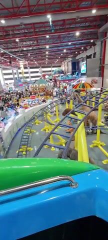 The video shows the perspective of a rider on a kiddie roller coaster in an indoor amusement park. The roller coaster car is blue and green. The track is yellow and blue.  Several animal figures and other attractions are seen as the ride progresses.  People can be seen in the background, including patrons and staff members. The park has a high ceiling with a red support structure.