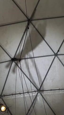 The video shows the inside of a dome structure with a white, textured inner surface supported by a gray metal frame. A shadow of a person performing aerial acrobatics on a hoop is projected onto the dome's surface. Several black ropes are suspended from the center of the dome, and a small light fixture is visible in the lower left corner.